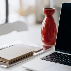 A minimalist home office setup featuring a laptop, notebook, and a red vase on a white desk.